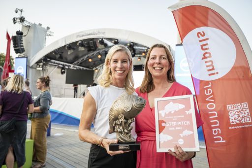 Zwei Frauen stehen vor einer Bühne. Eine hält eine Trophäe, die andere ein Schild mit „Bester Pitch".
