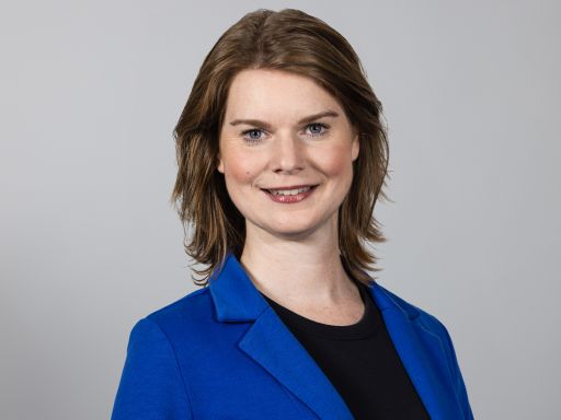 Frau mit schulterlangem braunem Haar, blauem Blazer, schwarzem Oberteil, lächelt vor grauem Hintergrund.
