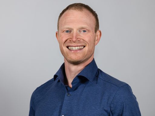 Mann mit roten Haaren und blauem Hemd lächelt vor grauem Hintergrund. Professionelles Porträtfoto.