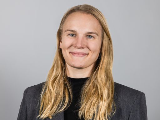 Porträtfoto einer Frau mit langen blonden Haaren, grauem Blazer und schwarzem Oberteil vor grauem Hintergrund.