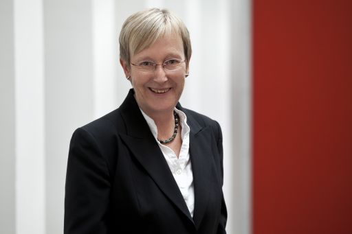 Lächelnde Frau mit kurzen blonden Haaren, Brille, schwarzem Blazer und Perlenkette vor weißem und rotem Hintergrund.