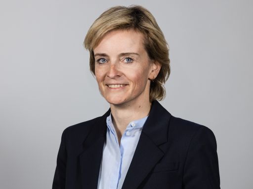 Frau mit blonden Haaren und blauen Augen in dunklem Blazer und hellblauem Hemd vor grauem Hintergrund.
