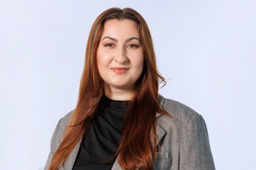 Frau mit langen roten Haaren, schwarzem Rollkragen und grauem Blazer vor hellgrauem Hintergrund.