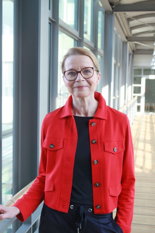 Frau mit Brille und roter Jacke über schwarzem Oberteil steht in einem Gebäude mit großen Fensterfronten.