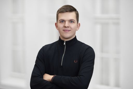 Junger Mann mit kurzem Haar, verschränkten Armen, schwarzem Zip-Pullover (Fred Perry), vor weißem Hintergrund.