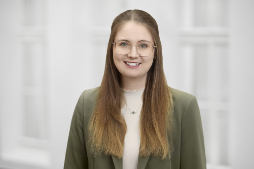 Junge Frau mit langen braunen Haaren und Brille, grünem Blazer, lächelt vor hellem Hintergrund.