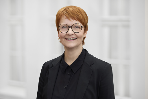 Frau mit rotem Kurzhaar und Hornbrille, schwarzem Blazer, lächelt vor hellem Hintergrund.