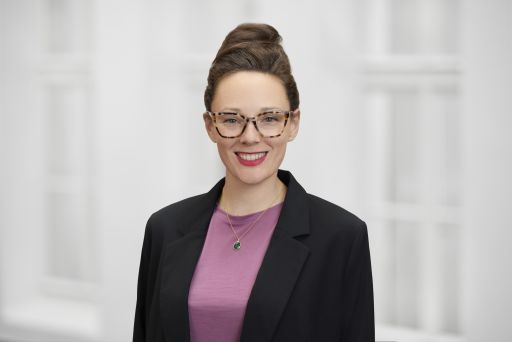 Frau mit Dutt, Brille, rotem Lippenstift, schwarzem Blazer, lila Shirt und grünem Anhänger lächelt vor weißem Hintergrund.