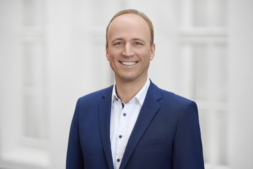 Mann in blauem Blazer und weißem Hemd lächelt vor hellem Hintergrund. Kurze blonde Haare, professionelles Porträtfoto.