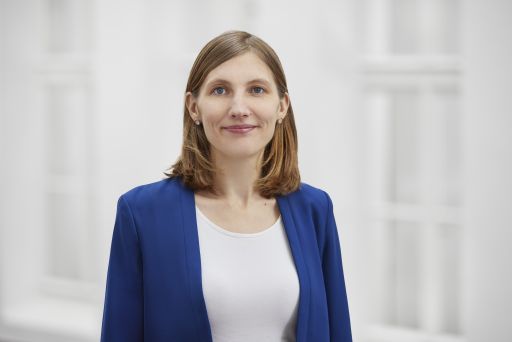 Frau mit schulterlangem braunem Haar, blauem Blazer und weißem Top, lächelnd vor hellem Hintergrund.