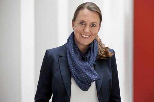 Lächelnde Frau mit Pferdeschwanz, blauem Blazer und gemustertem Schal vor hellem Hintergrund.