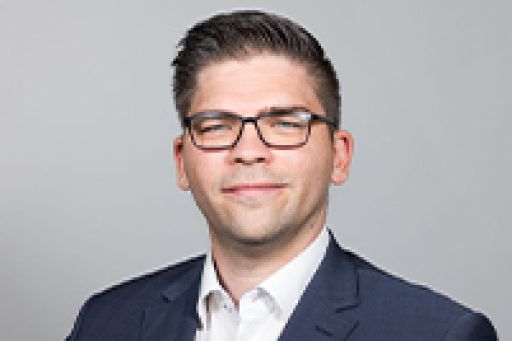 Mann mit Brille und dunklem Sakko, weißem Hemd, vor grauem Hintergrund, professionelles Porträtfoto.