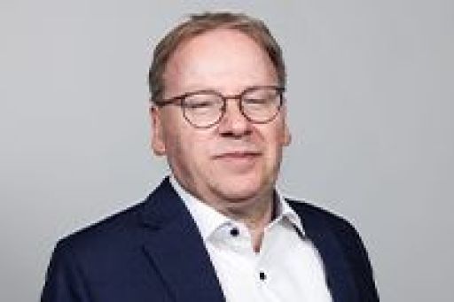 Mann mittleren Alters mit Brille und blondem Haar, trägt blaues Jackett, vor grauem Hintergrund.