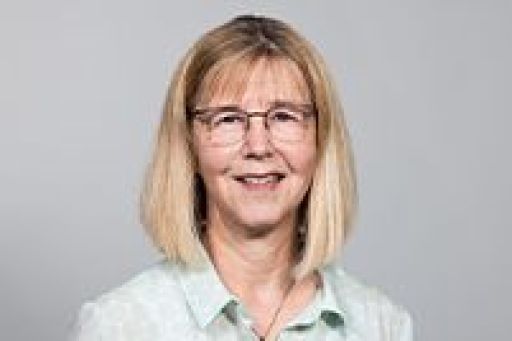 Frau mit blondem Bob, Brille und hellgrüner Bluse lächelt vor grauem Hintergrund.