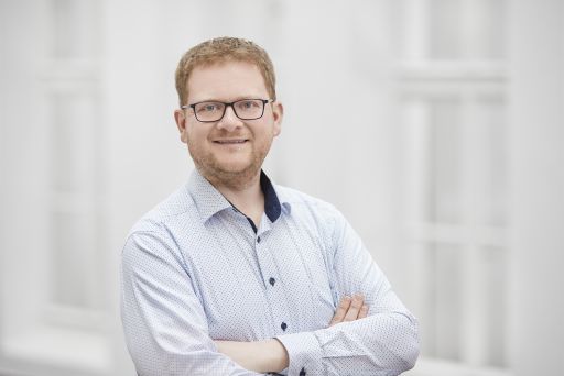Mann mit rotem Bart und Brille, hellblaues Hemd, Arme verschränkt, lächelnd vor hellem Hintergrund.