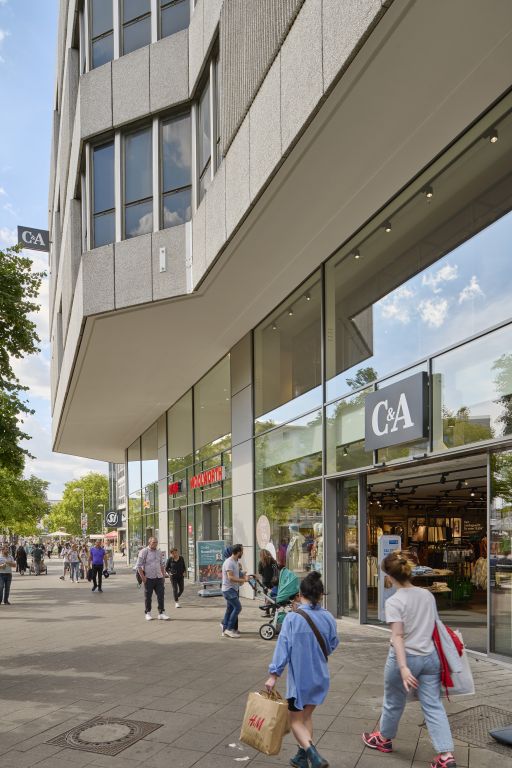 Fußgängerzone mit C&A-Filiale im Erdgeschoss eines modernen Hochhauses, Passanten mit Kinderwagen und H&M-Einkaufstasche.