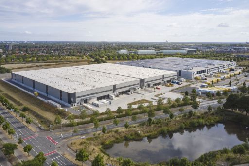 Luftaufnahme eines großen Logistikzentrums mit Flachdach, Parkplätzen und Laderampen, umgeben von Grünflächen und Straßen.