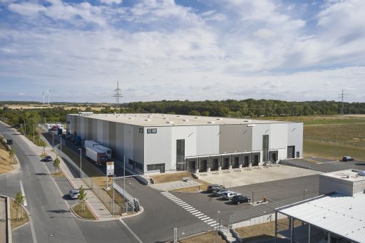 Luftaufnahme eines großen Industriegebäudes mit Lkw-Rampen, Parkplatz, Straße, Windrädern und Hochspannungsmasten im Hintergrund.