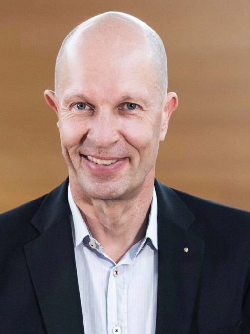 Kahler Mann mittleren Alters in dunklem Sakko und hellblauem Hemd lächelt vor braunem Hintergrund.
