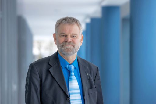 Mann mit grauem Bart und Haar, blauem Hemd, gestreifter Krawatte und dunklem Sakko steht in einem Flur.