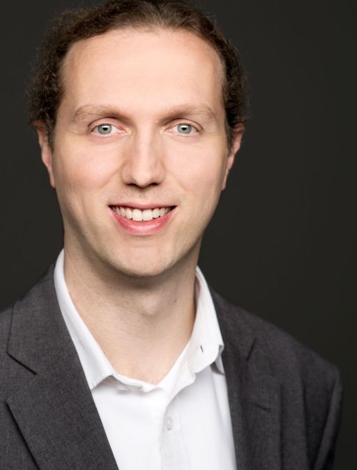 Mann mit kurzen braunen Haaren und blauen Augen, weißem Poloshirt und grauem Sakko, lächelt vor dunklem Hintergrund.
