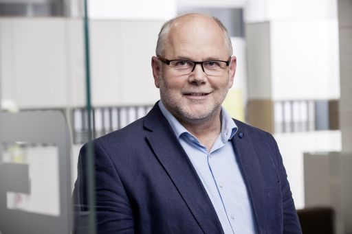 Mann mit Brille und Glatze in blauem Sakko lächelt vor unscharfem Büro-Hintergrund mit Aktenordnern.