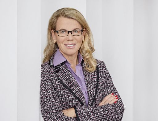 Frau mit blonden Haaren und Brille, lila Bluse, kariertem Blazer, verschränkte Arme, lächelt in die Kamera.