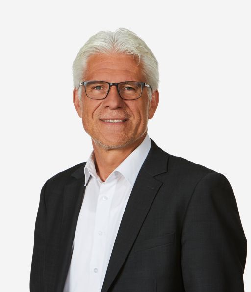 Älterer Mann mit weißem Haar und Brille, in dunklem Sakko und weißem Hemd, lächelt in die Kamera.