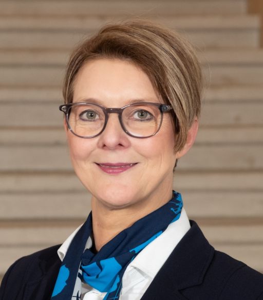 Frau mit kurzen blonden Haaren und Brille, blauem Blazer und blauem Schal, lächelt in die Kamera.