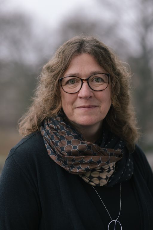 Frau mit lockigem Haar, Brille und gemustertem Schal, trägt schwarze Jacke und Kette, vor unscharfem Hintergrund.