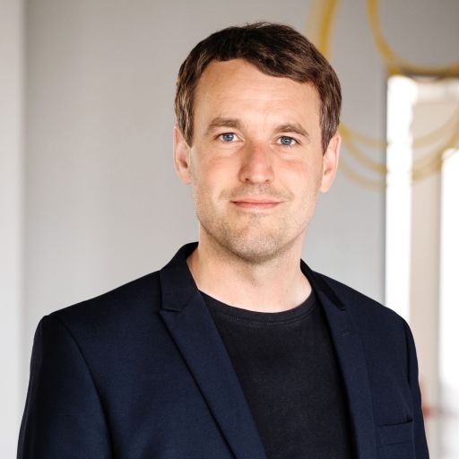 Mann mit braunen Haaren und blauen Augen, dunklem Blazer über schwarzem T-Shirt, vor hellem Hintergrund.