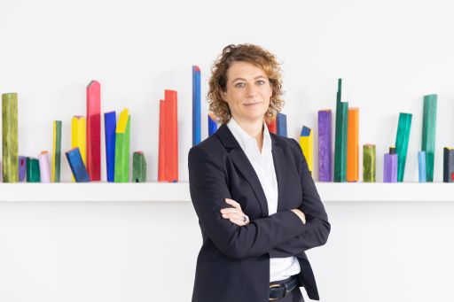 Frau mit lockigem Haar und schwarzem Blazer steht mit verschränkten Armen vor bunten Holzstäben an weißer Wand.