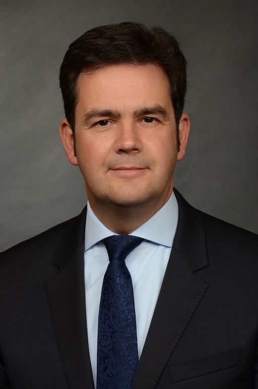Mann in dunklem Anzug mit hellblauem Hemd und dunkelblauer Krawatte vor grauem Hintergrund. Professionelles Porträtfoto.