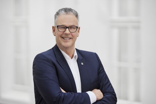 Mann mit Brille und grauem Haar, dunklem Blazer, Arme verschränkt, lächelt, heller Hintergrund.