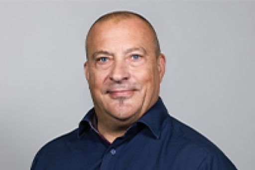 Mann mittleren Alters mit kurzem Haar, dunklem Hemd, lächelt vor grauem Hintergrund.