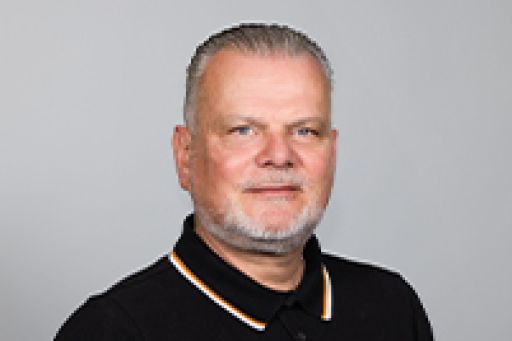 Älterer Mann mit grauen Haaren, schwarzem Polo-Shirt mit gelbem Streifen, vor grauem Hintergrund.