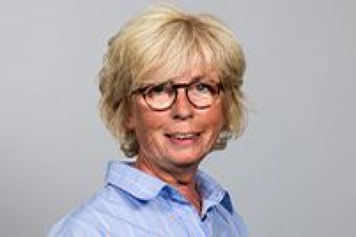 Lächelnde Frau mit blondem Kurzhaar und Brille in hellblauem Hemd vor grauem Hintergrund.
