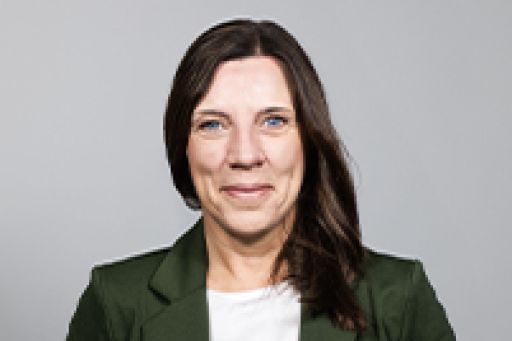 Frau mit dunkelbraunem Haar, grüner Jacke und weißem Oberteil lächelt vor grauem Hintergrund.