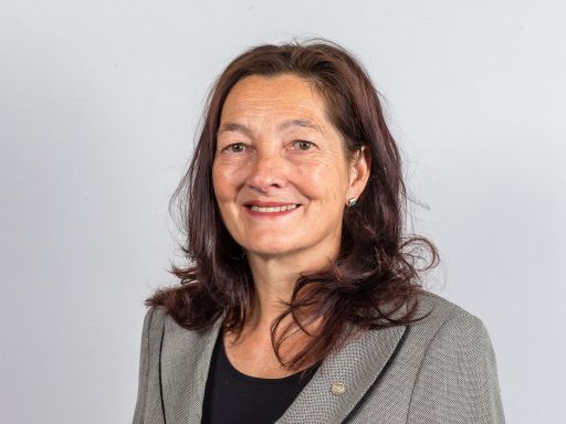 Frau mit dunkelrotem Haar, grauem Blazer und schwarzem Oberteil lächelt vor hellgrauem Hintergrund.