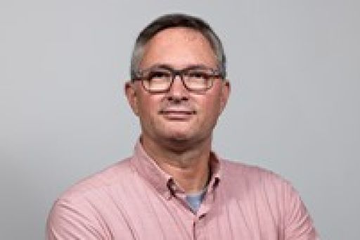 Mann mittleren Alters mit Brille und kurzem grauem Haar, rosa Hemd, vor grauem Hintergrund.