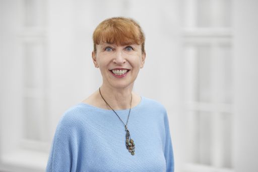 Lächelnde Frau mit roten kurzen Haaren, blauen Augen, hellblauem Pullover und einer Anhängerkette vor weißem Hintergrund.