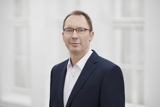 Mann mit Brille und Blazer, weißes Hemd, vor hellem Hintergrund, Porträtaufnahme.
