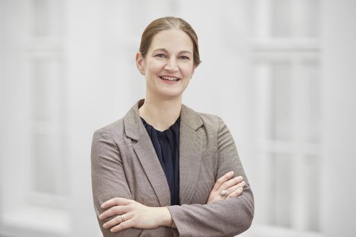 Frau mit zurückgebundenem Haar, beigem Blazer und blauer Bluse, Arme verschränkt, lächelt in die Kamera.