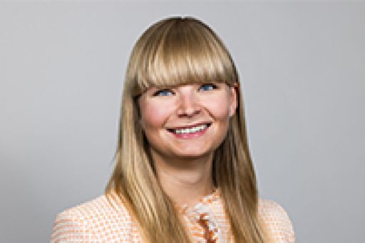 Frau mit langen blonden Haaren und Pony, lächelnd, trägt ein hellrosa Oberteil, vor grauem Hintergrund.