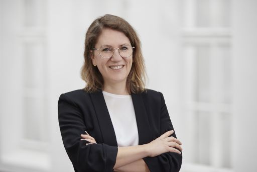 Frau mit Brille und schulterlangen Haaren, schwarzem Blazer, Arme verschränkt, lächelt in die Kamera.