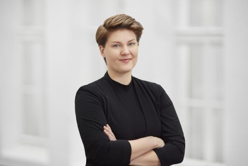 Person mit kurzem Haar in schwarzem Oberteil, Arme verschränkt, vor hellem Hintergrund, lächelnd.