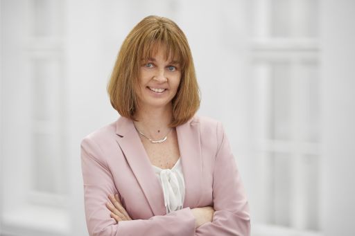 Frau mit rötlich-braunem Bob, rosa Blazer und Perlenkette, Arme verschränkt, lächelt vor hellem Hintergrund.