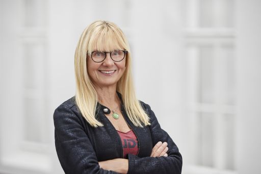 Lächelnde Frau mit blonden Haaren und Brille, verschränkten Armen, dunklem Blazer, vor hellem Hintergrund.