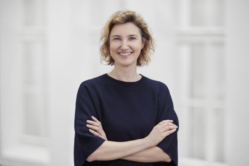 Lächelnde Frau mit kurzen blonden Haaren, dunklem Pullover, verschränkten Armen, vor hellem Hintergrund.