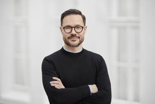 Mann mit Brille und Bart, schwarzem Pullover, Arme verschränkt, lächelt, heller Hintergrund.